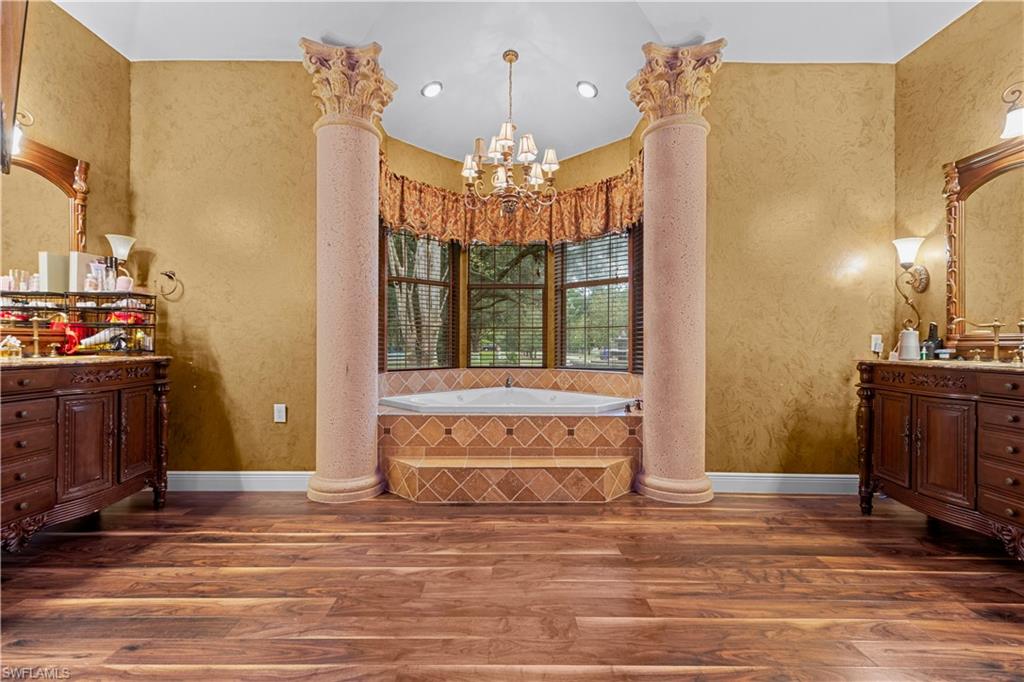 Spacious bathroom featuring a built-in jetted tub with tiled surround, two decorative columns, a chandelier, and wood-finish flooring