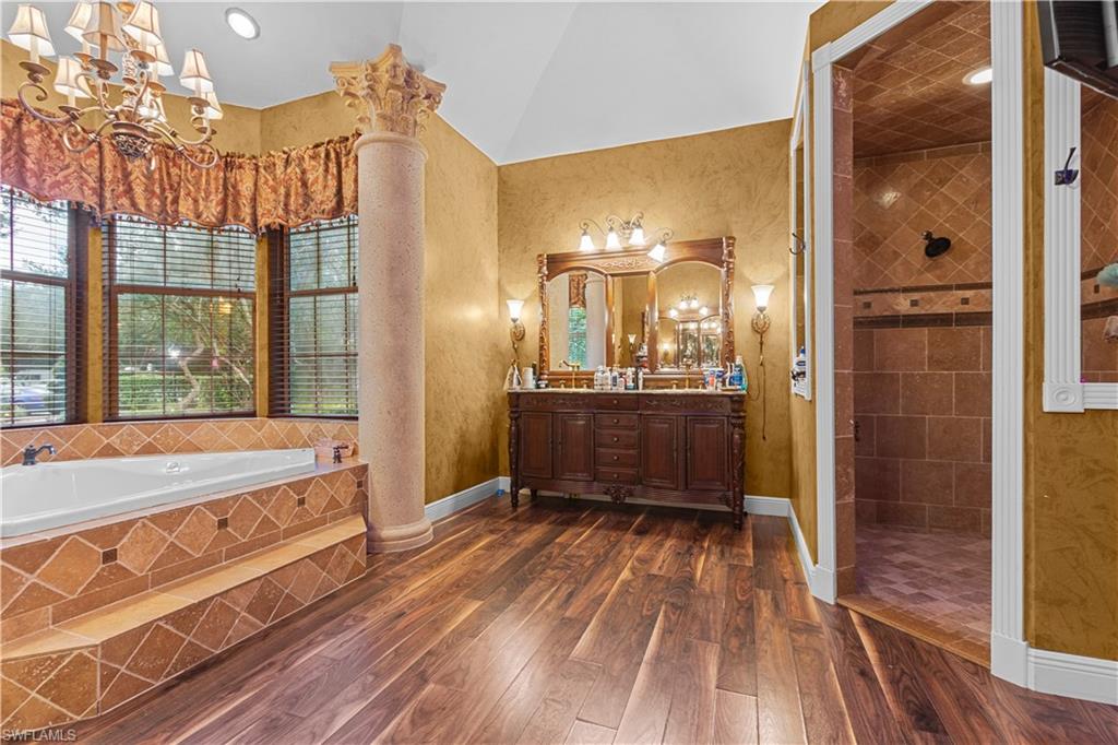 Spacious bathroom featuring a large soaking tub with tiled surround, an ornate column, a double vanity with framed mirrors, a walk-in shower with tiled walls and flooring, and wood-finish flooring