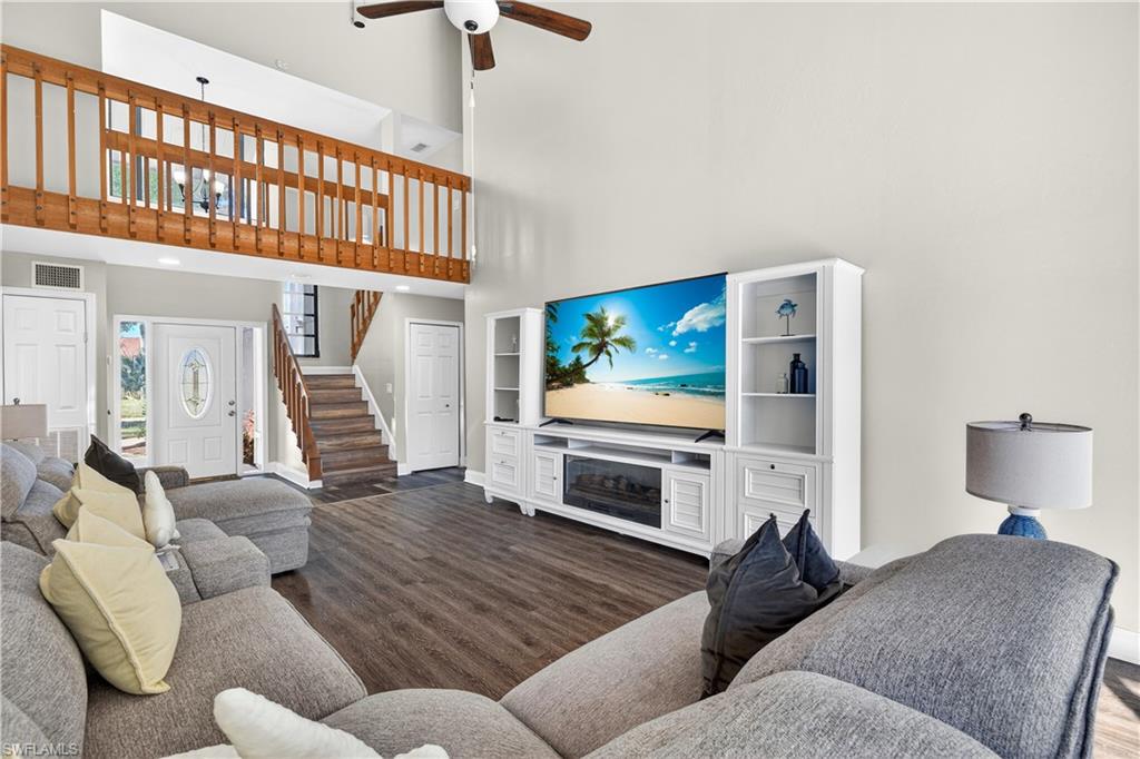 Vaulted living space featuring wood-finish flooring, a white built-in media console with an electric fireplace, a wood-railed loft overlook, a matching wood staircase, and a front entry door with decorative glass