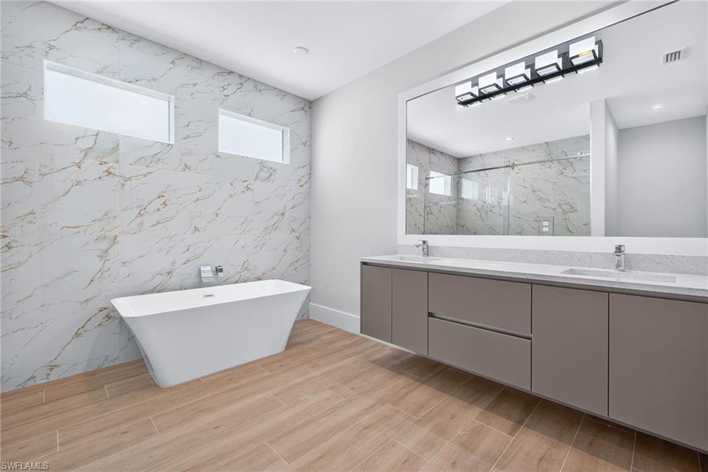 Bathroom featuring a freestanding soaking tub, large format wall tiling, wood-finish floor tiling, dual vanity with undermount sinks, and a frameless mirror