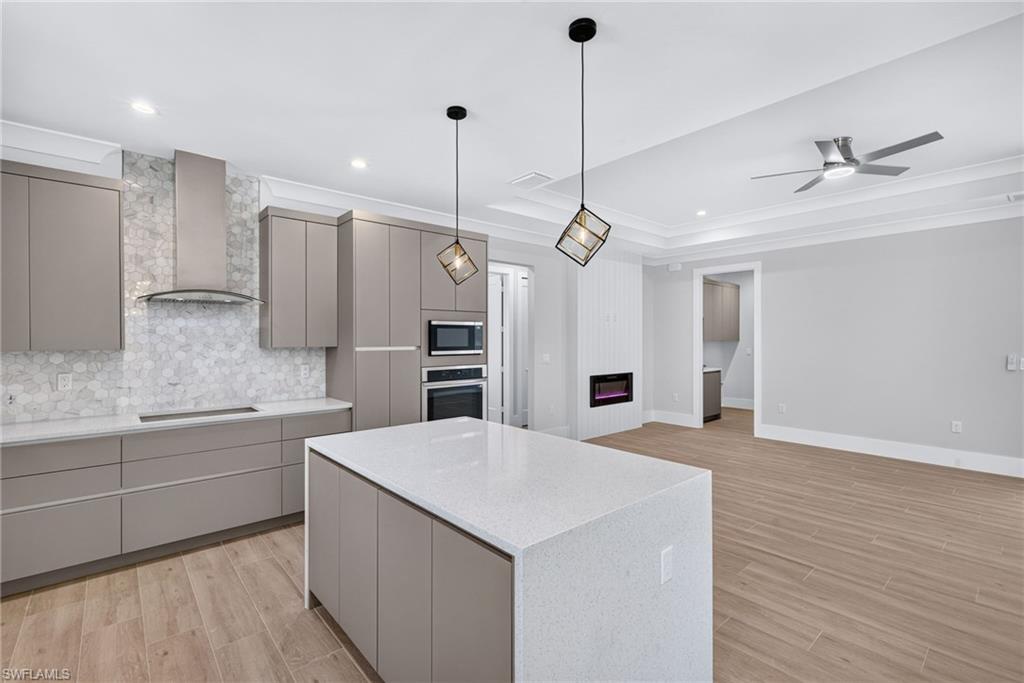 Open-concept kitchen featuring an oversized island with a solid surface countertop, matte gray cabinetry, a hexagonal tile backsplash, stainless steel appliances, and wood-finish flooring