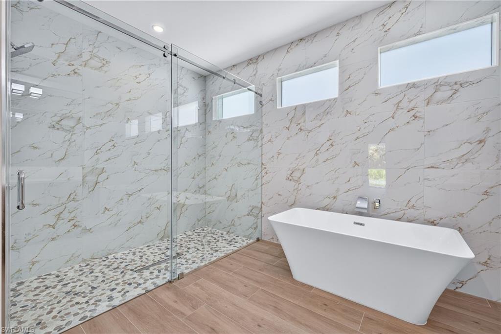 Spacious bathroom featuring a freestanding tub, large glass-enclosed shower with pebble floor, wood-finish flooring, and wall-mounted windows