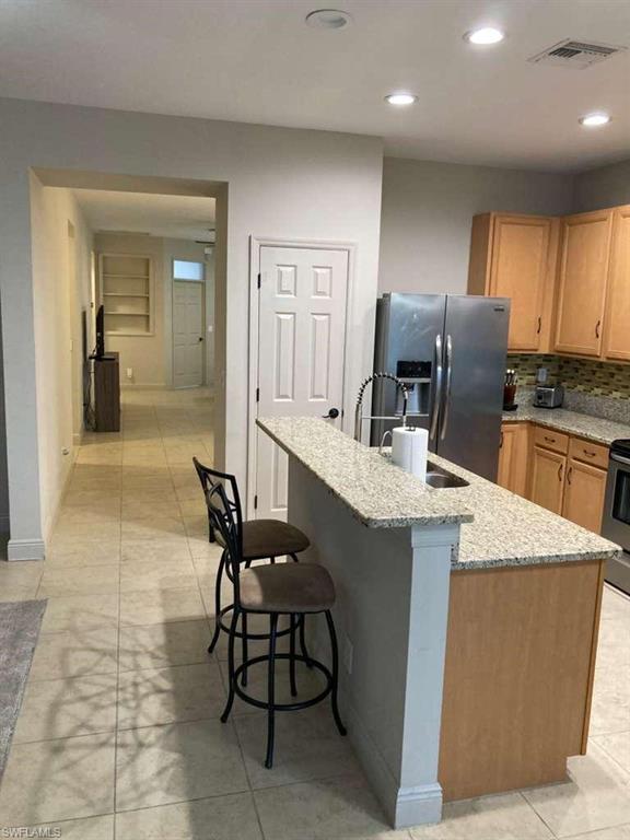 Kitchen featuring a granite-topped island with breakfast bar seating, light wood-finish cabinetry, stainless steel appliances, and tile flooring