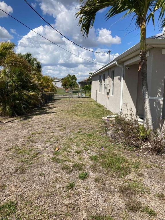 Side yard featuring a chain-link fence, mature palm trees, and established shrubs bordering the stucco exterior