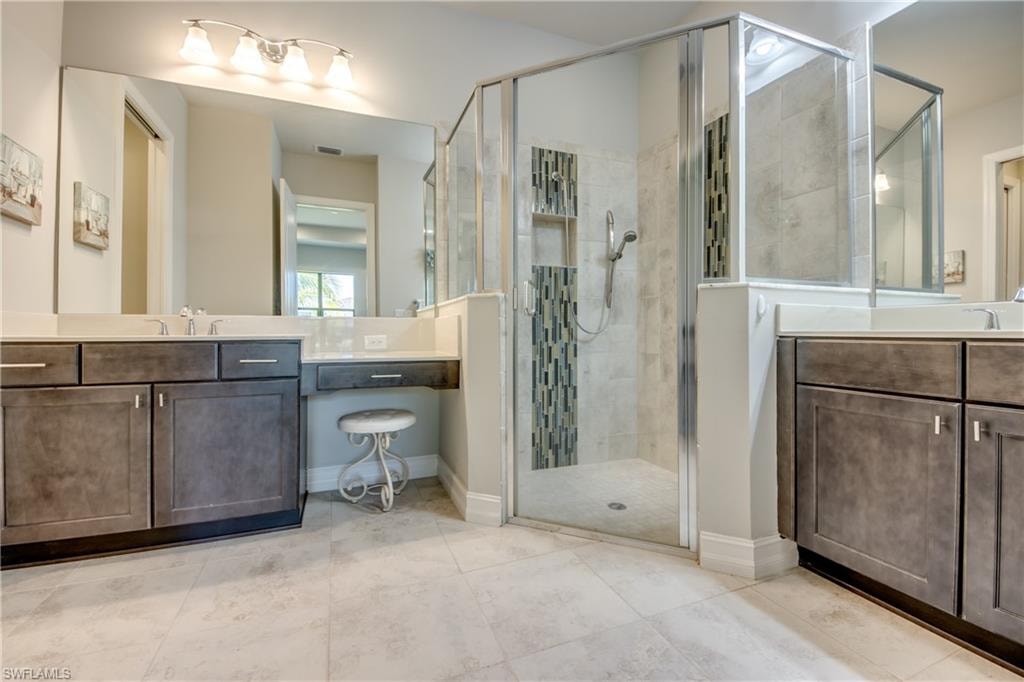 Spacious bathroom featuring a glass-enclosed shower with decorative tile accent, dual vanity sinks, a dedicated makeup counter, and large format floor tiling
