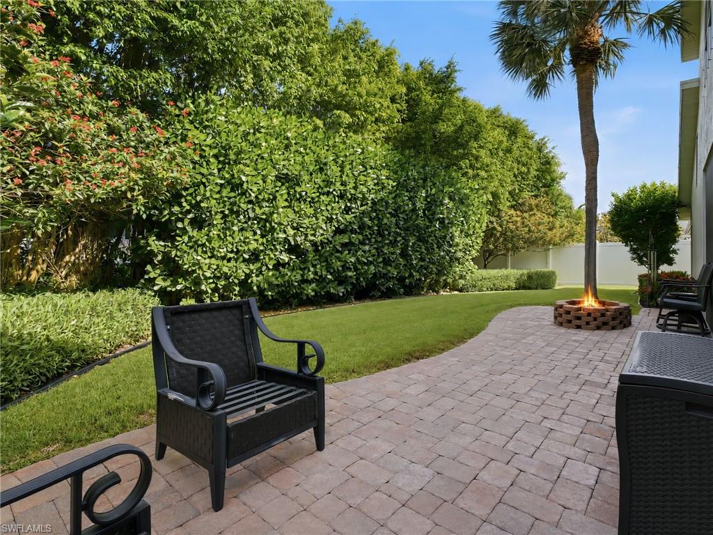 Expansive paved patio featuring a built-in fire pit, surrounded by lush green landscaping and mature trees