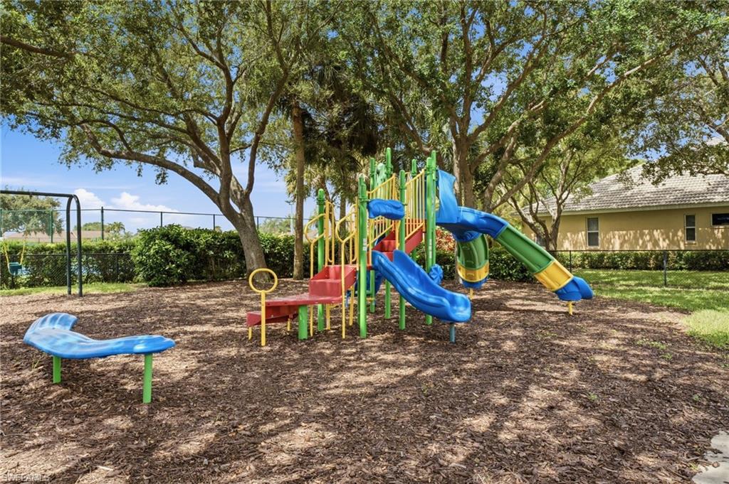 Outdoor recreational area featuring a multi-color play structure with slides and climbing elements, a swing set, mature shade trees, and a ground cover of wood chips