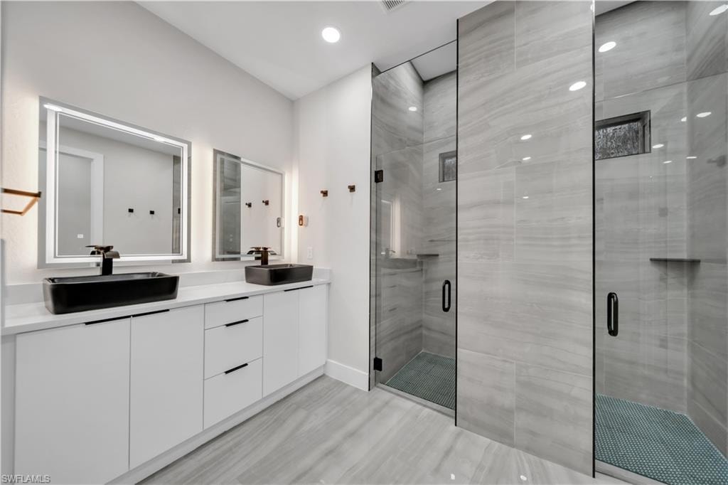 Dual shower stalls with glass enclosures, an elongated double vanity, backlit mirrors, black vessel sinks, and wood-finish flooring