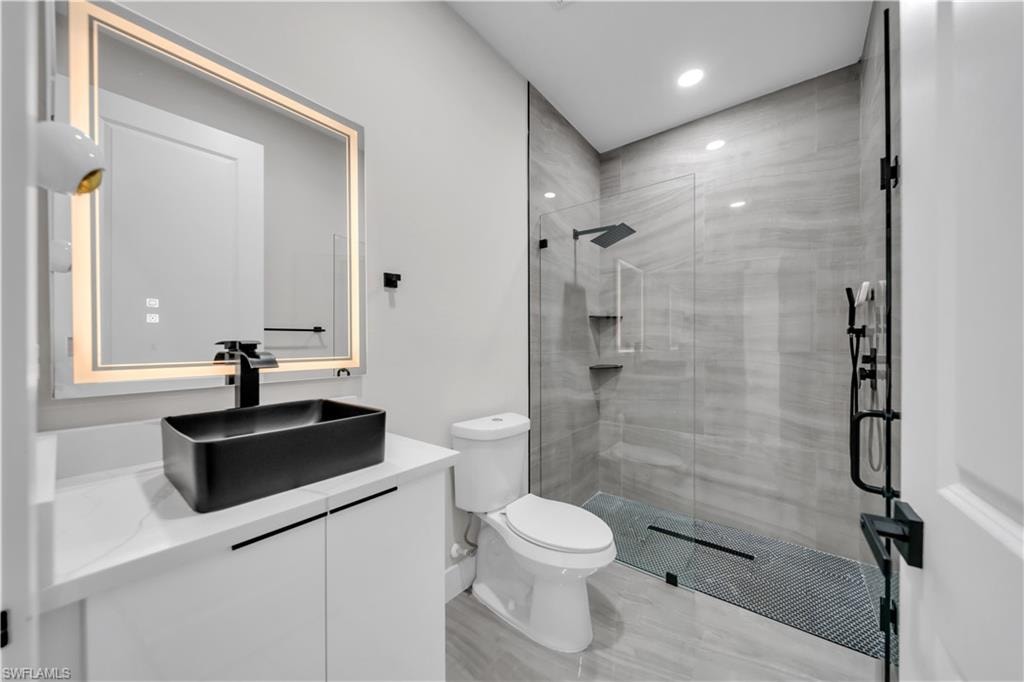 Rectangular vessel sink with matte black faucet, illuminated mirror, white vanity with concealed hardware, glass-enclosed shower, and large format wall tiling