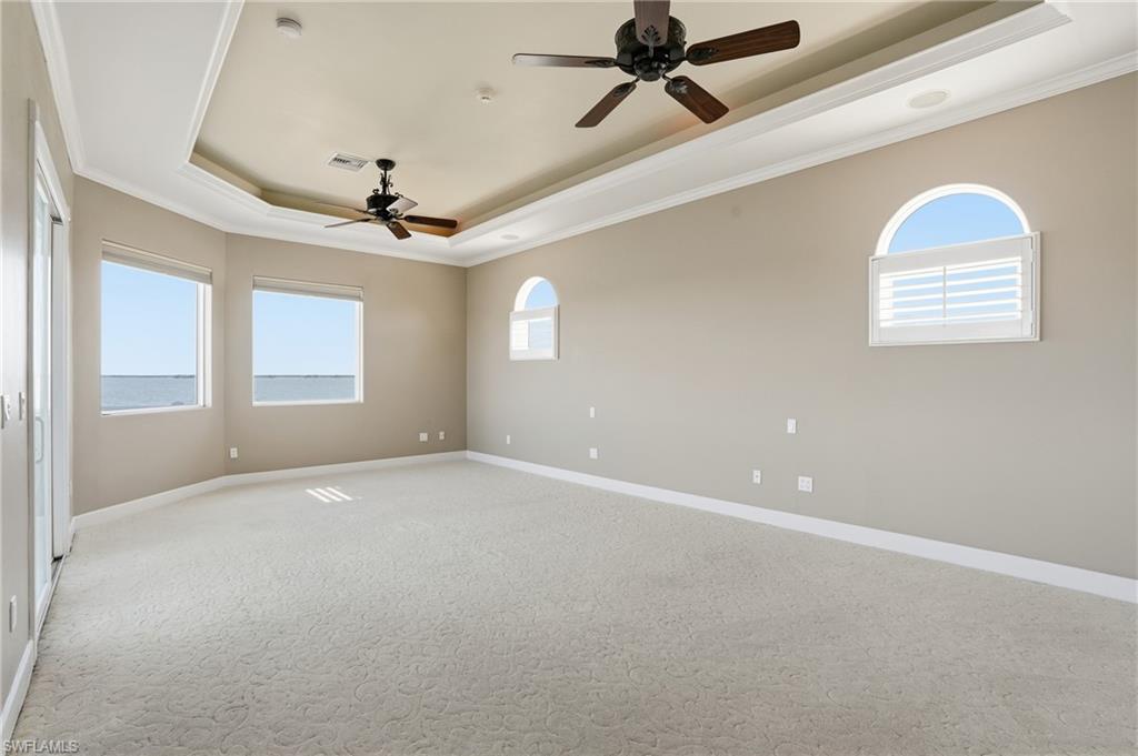 Unfurnished room featuring ornamental molding, a raised ceiling, light colored carpet, ceiling fan, and plenty of natural light - Virtually Edited Image