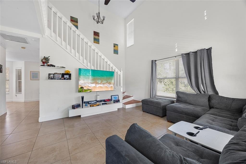 Living area featuring high ceilings, tile flooring, a prominent staircase with white railings, a large window with blinds, and a ceiling-mounted light fixture