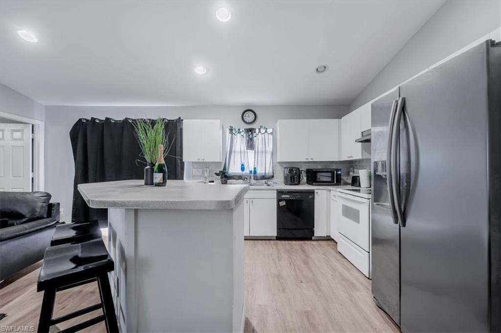 Kitchen featuring a central peninsula with a light-toned countertop, white cabinetry, a stainless steel refrigerator, black and white appliances, and wood-finish flooring