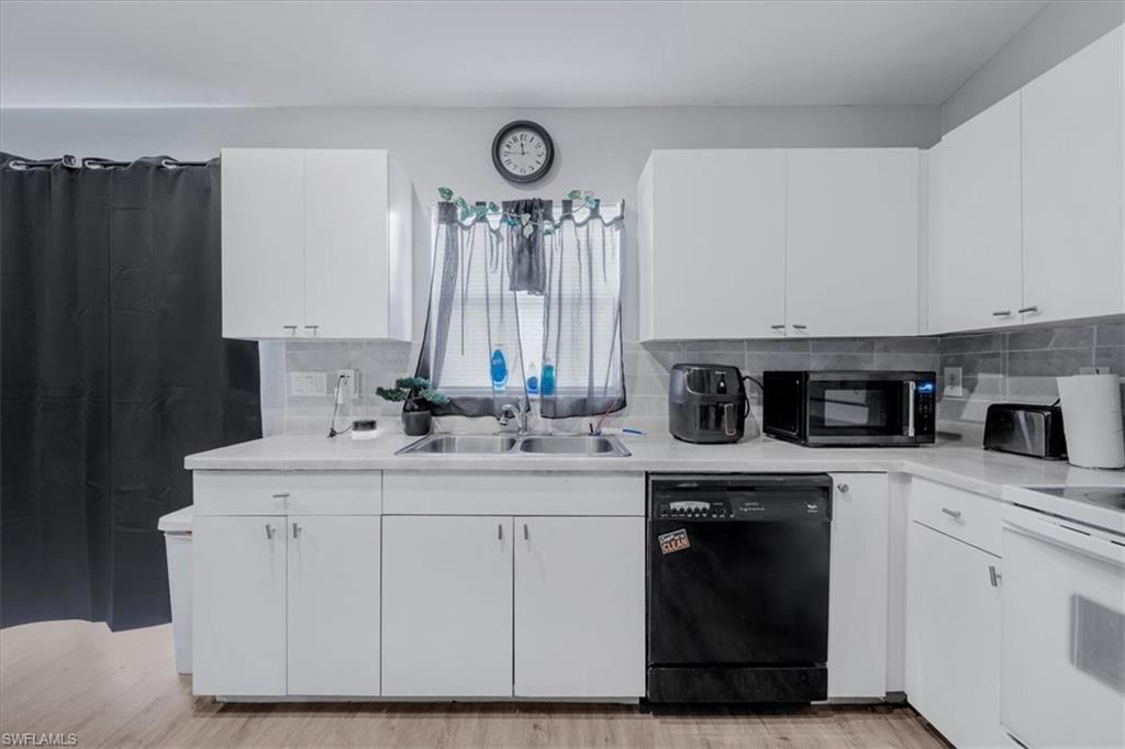 Kitchen featuring white cabinetry, light-toned countertops, a stainless steel double sink, a dark tile backsplash, and wood-finish flooring