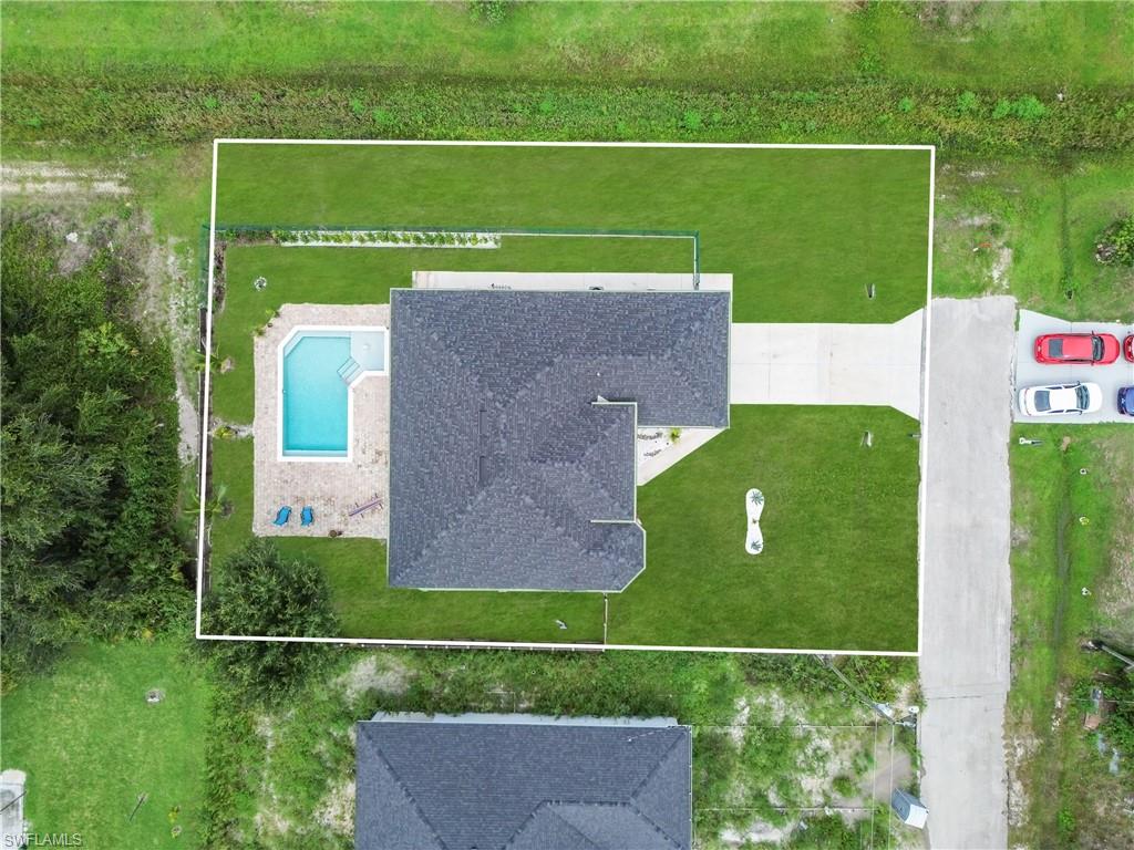 Aerial view featuring a residential property with a dark shingle roof, a geometric swimming pool with paved decking, and a concrete driveway