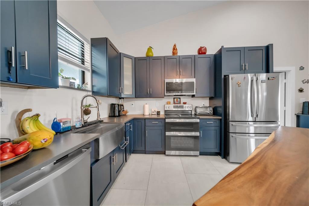 Modern kitchen featuring blue cabinetry, stainless steel appliances, a large farmhouse sink, white tile flooring, and a vaulted ceiling