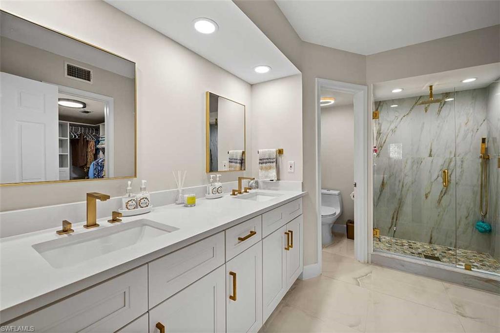 Contemporary bathroom featuring a dual vanity with white cabinetry and integrated sinks, brushed gold fixtures, a spacious walk-in shower with a glass enclosure and marble-look wall tiles, and recessed lighting