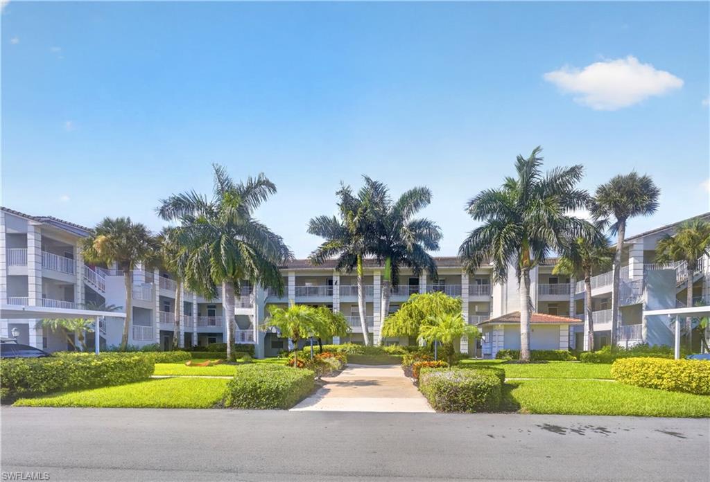 Multi-story building with multiple balconies, mature palm trees, manicured landscaping, paved pathways, and covered parking structures
