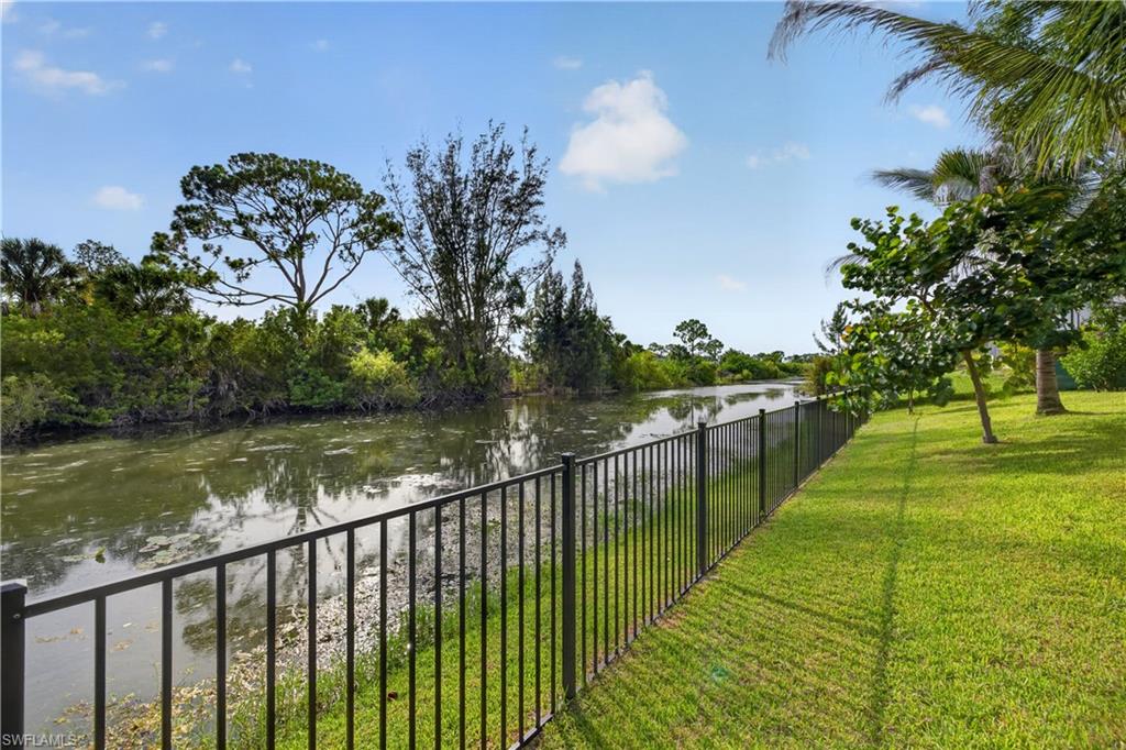 Expansive waterside property featuring a black metal fence, lush green lawn, and mature tropical landscaping