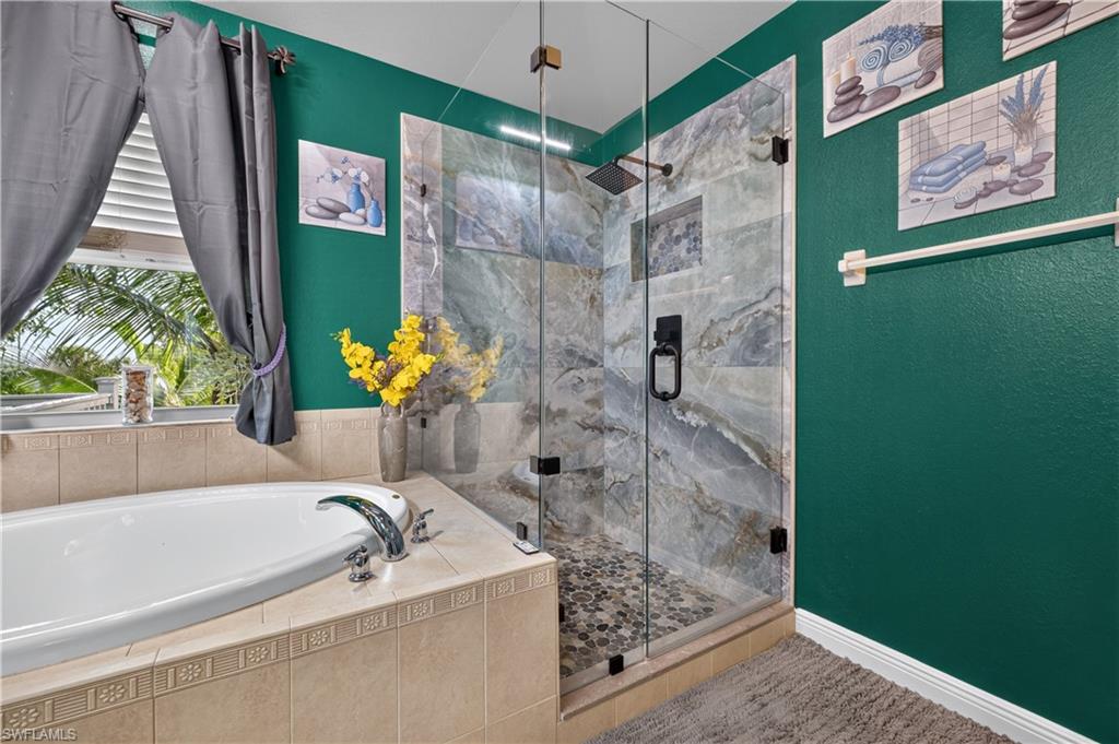 Bathroom featuring a glass-enclosed shower with pebble flooring and stone-look wall tiles, a built-in tub with chrome fixtures, and a window with horizontal blinds and privacy drapes