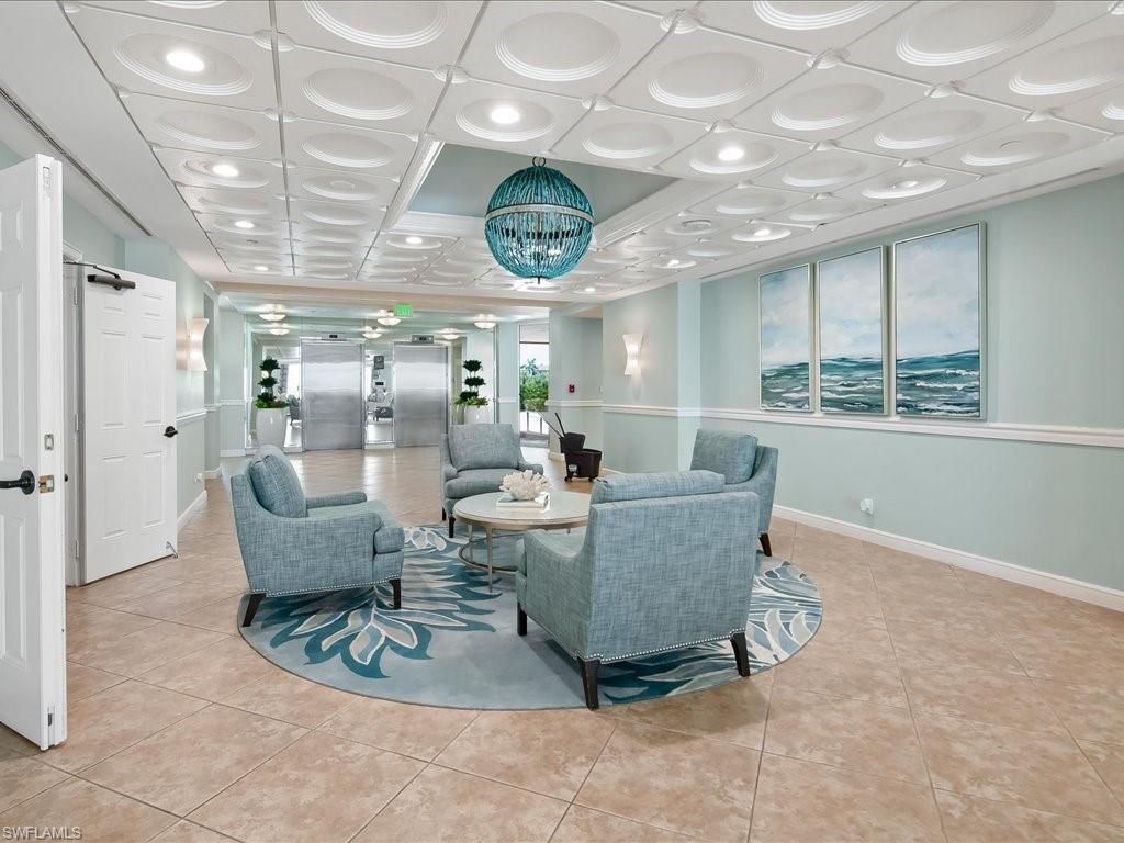 Lobby lounge featuring patterned ceiling tiles with recessed lighting, blue spherical chandelier, four armchairs surrounding a round coffee table, and large format floor tiling