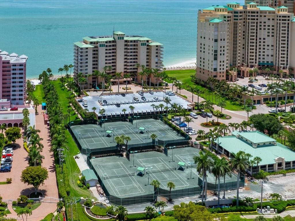 Community tennis courts with multiple playing surfaces, surrounded by manicured landscaping and mature palm trees, offering proximity to beachfront high-rise residences and the ocean