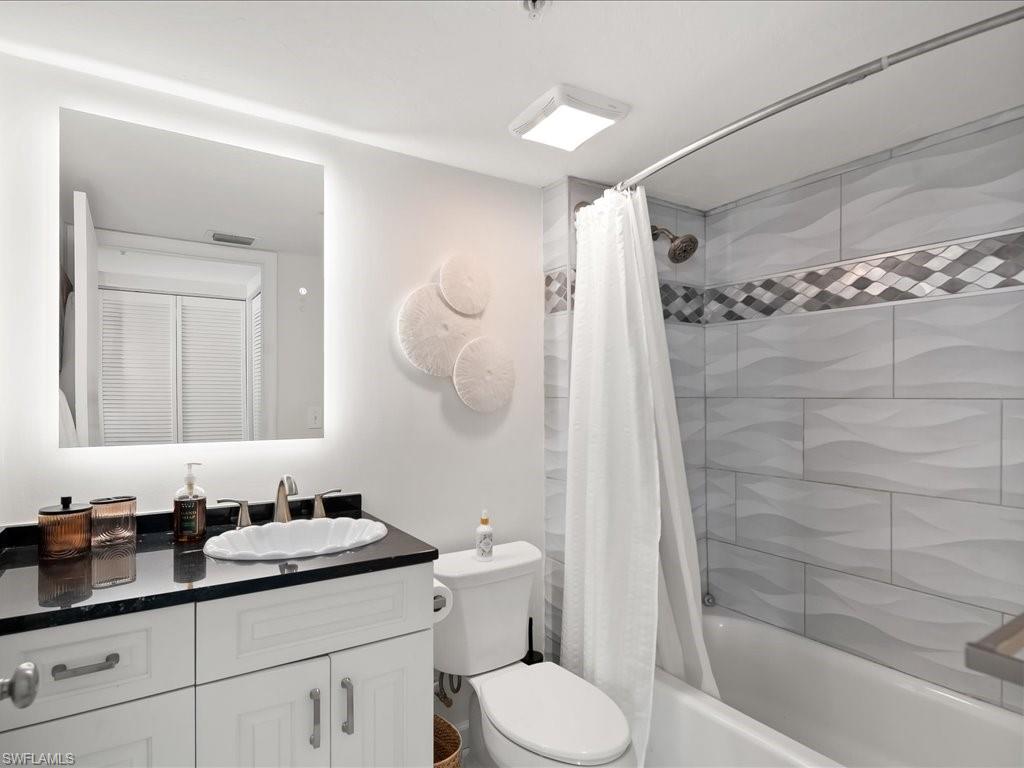 Bathroom featuring a white vanity with dark countertop, white shell-shaped sink, illuminated mirror, and a tiled shower with decorative accent band