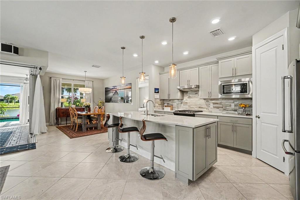 Contemporary kitchen featuring light gray cabinetry, a white countertop island with pendant lighting, a tile backsplash, stainless steel appliances, and large format tile flooring