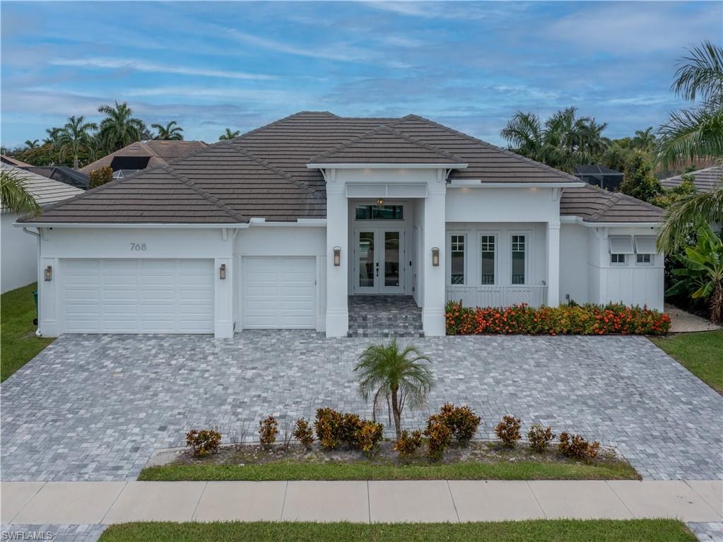 Modern residence featuring a striking pitched roof with dark tile detailing, clean white exterior with architectural pillars framing the entrance, triple-car garage configuration, and a paver driveway with manicured landscaping including flowering shrubs and palm trees