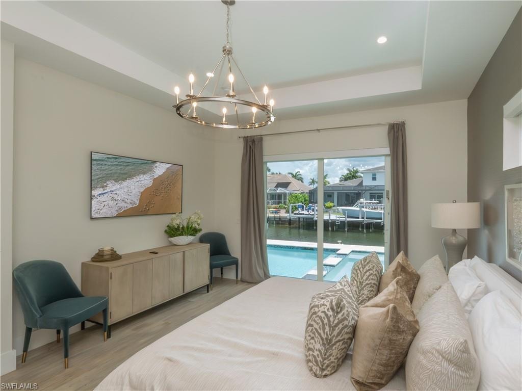 Master bedroom suite with waterfront access, featuring a wood-finish plank floor, soft-close cabinetry, pendant lighting fixture, floor-to-ceiling sliding glass doors opening to a pool and water views, and an ensuite sitting area with upholstered seating