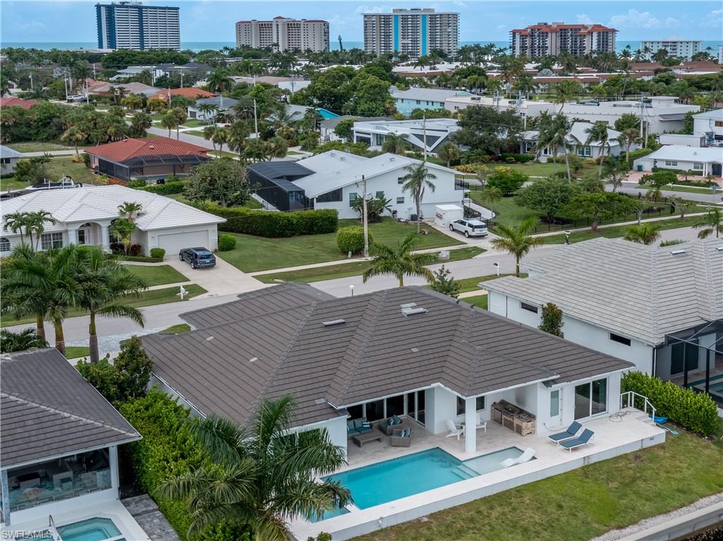 Modern waterfront residence showcasing a resort-style heated swimming pool, expansive lanai with integrated seating areas, metal roof with clean architectural lines, manicured lawn with mature tropical landscaping, and direct sightlines to the intracoastal waterway and high-rise developments beyond