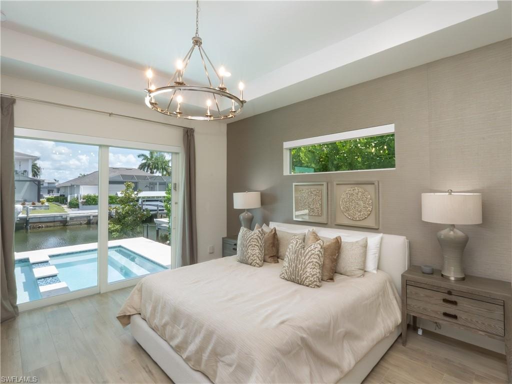 Primary Bedroom with Sliding Glass Doors to Pool Area, Light Wood-Finish Flooring, Neutral Textured Wall Finishes, Pendant Chandelier with Integrated Lighting, and Dual Nightstands with Built-In Storage
