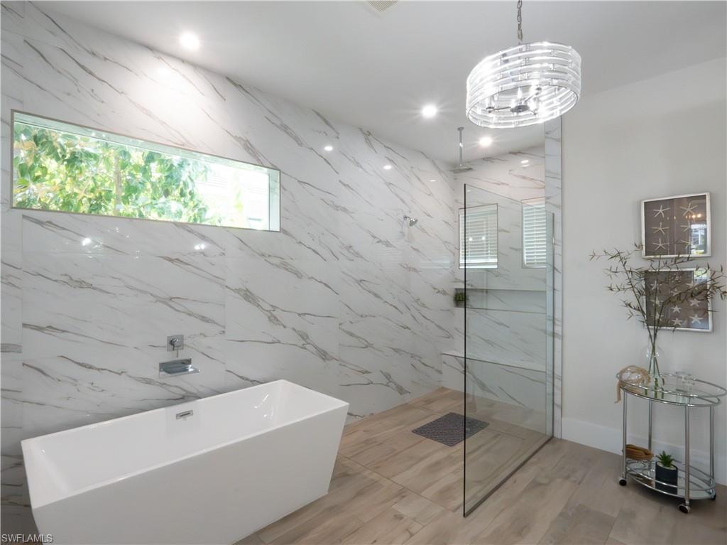 Freestanding soaking tub, walk-in shower with glass enclosure, marble-style wall tiling, wood-finish flooring, and a linear transom window