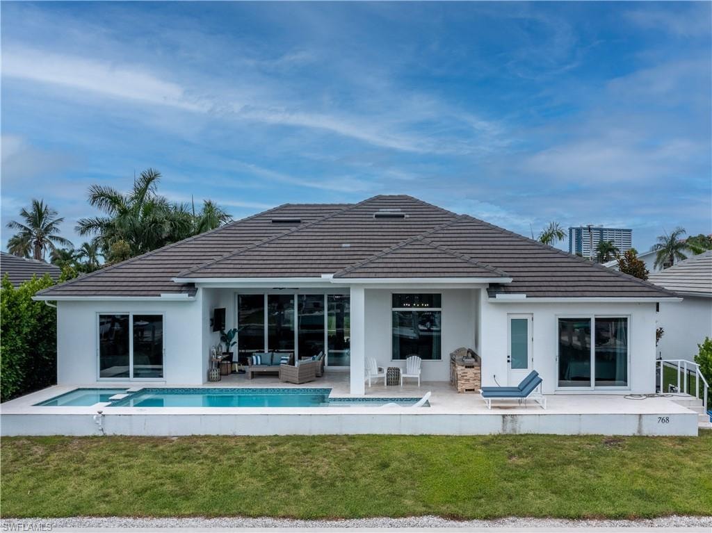 Modern architectural residence featuring a clean-lined white exterior with charcoal tile roofing, expansive glass sliding doors and windows, and a resort-style backyard with heated swimming pool, mature tropical landscaping including palm trees, manicured lawn, and integrated hardscape patio areas ideal for outdoor entertaining