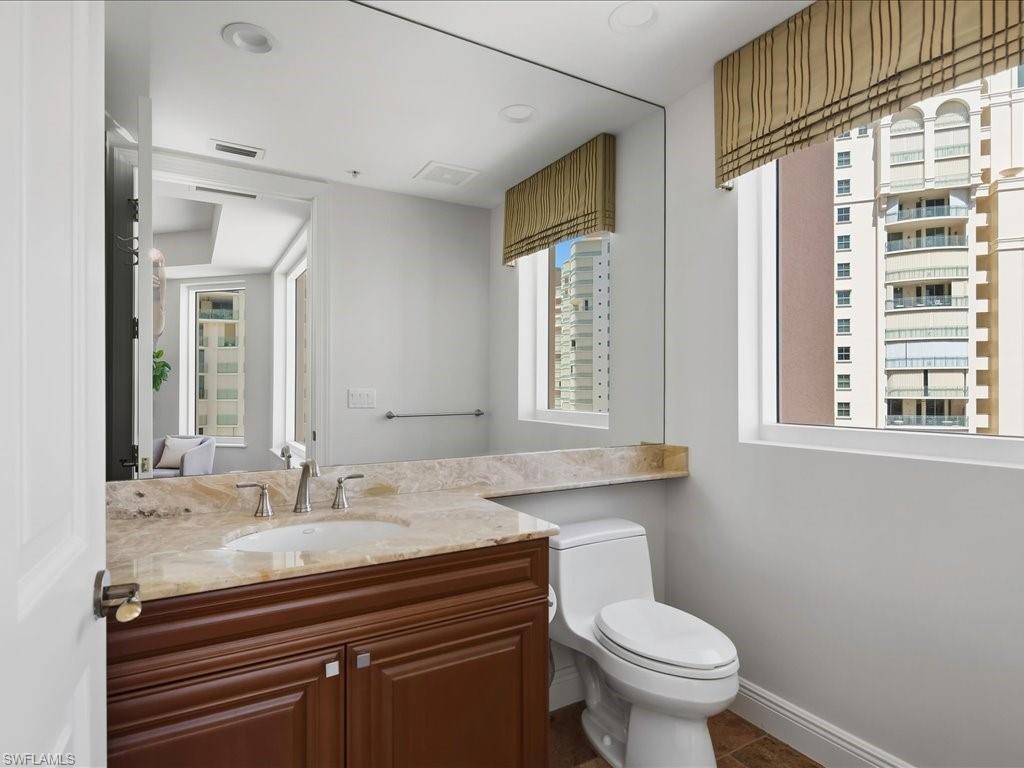 Bathroom featuring a built-in vanity with a stone countertop and integrated sink, a large wall-mounted mirror, a commode, and a window with a Roman shade