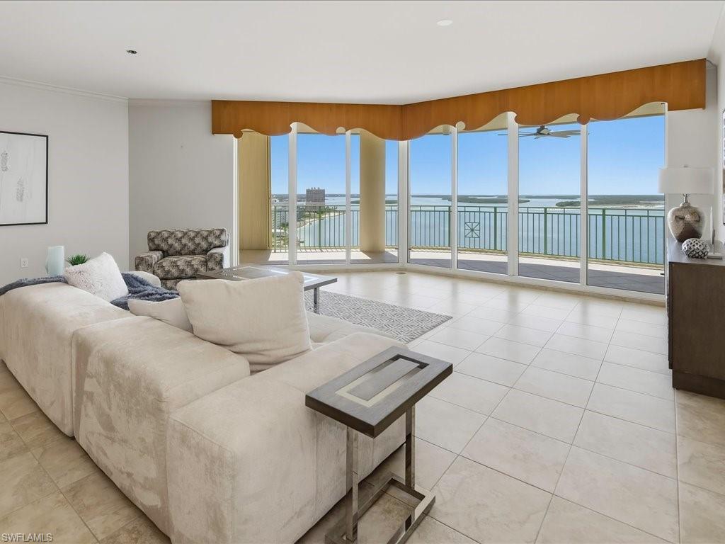 Bright living area featuring expansive floor-to-ceiling windows, light-colored tile flooring, a decorative wood valance, and access to a balcony with green railings overlooking a body of water