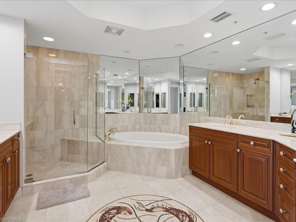 Spacious bathroom featuring a built-in soaking tub, a glass-enclosed shower, dual wood-finish vanities with light countertops, extensive wall-mounted mirrors, and tiled flooring with a decorative mosaic inlay