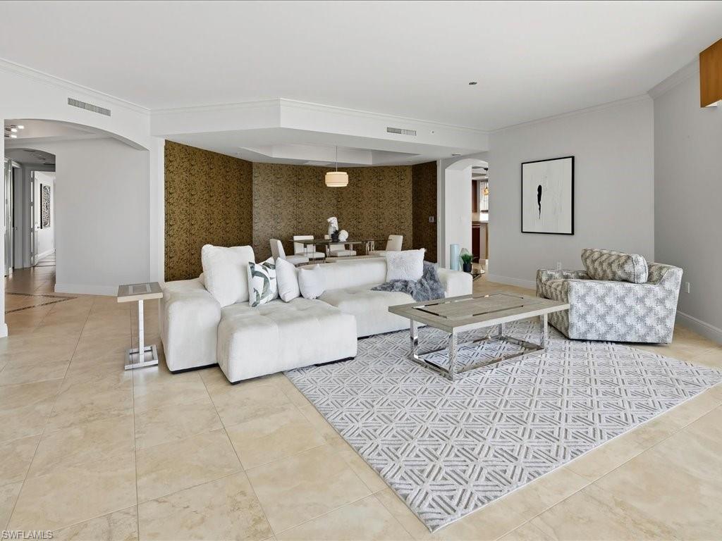 Spacious open-concept living area featuring light-toned tile flooring, crown molding, an arched entryway, and a dedicated dining space with distinctive wall covering
