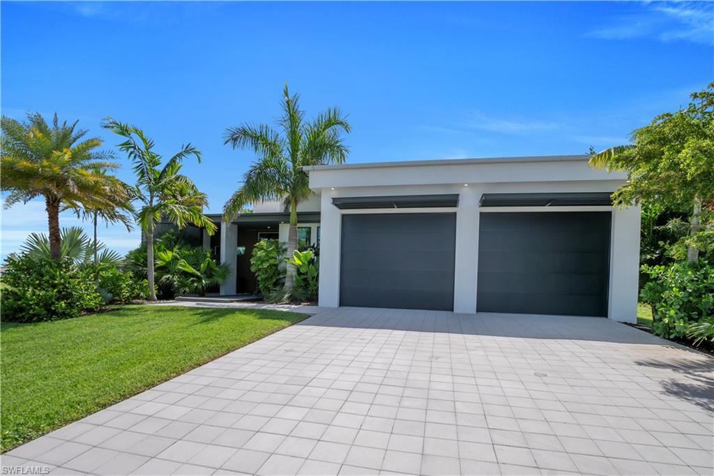 Modern exterior featuring a two-car garage with dark paneled doors, white stucco facade, a light-colored paver driveway, and a manicured lawn with tropical landscaping