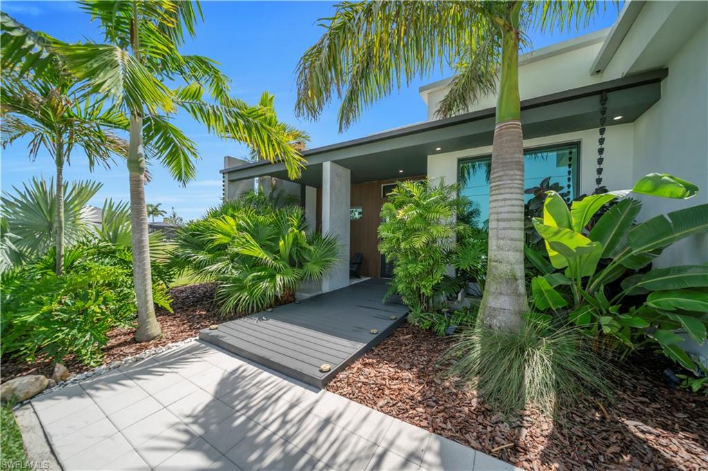Modern exterior with a light-colored facade, featuring lush tropical landscaping, a plank walkway, and a paver pathway
