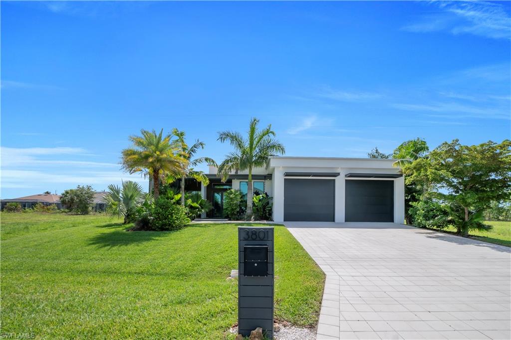 Modern architecture featuring a two-car garage, lush landscaping with palm trees, a paver driveway, and a manicured lawn