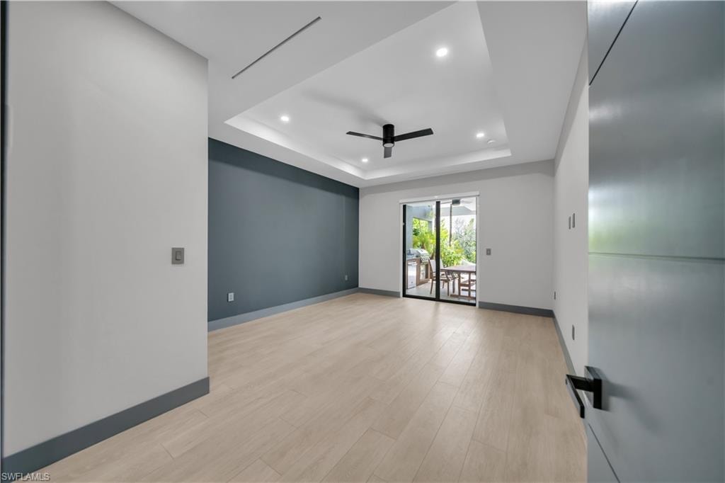 Spacious interior with light wood-finish flooring, a tray ceiling featuring recessed lighting and a ceiling fan, a contrasting accent wall, and sliding glass doors opening to an outdoor patio with a dining area
