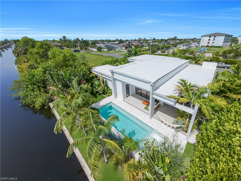 Modern waterside residence featuring a rectangular pool, covered patio, lush tropical landscaping, and a flat roofline