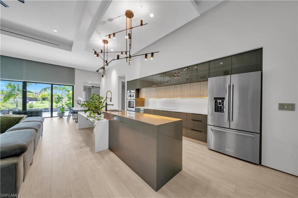 Modern kitchen featuring a large waterfall island, stainless steel refrigerator, integrated double ovens, and a contemporary chandelier