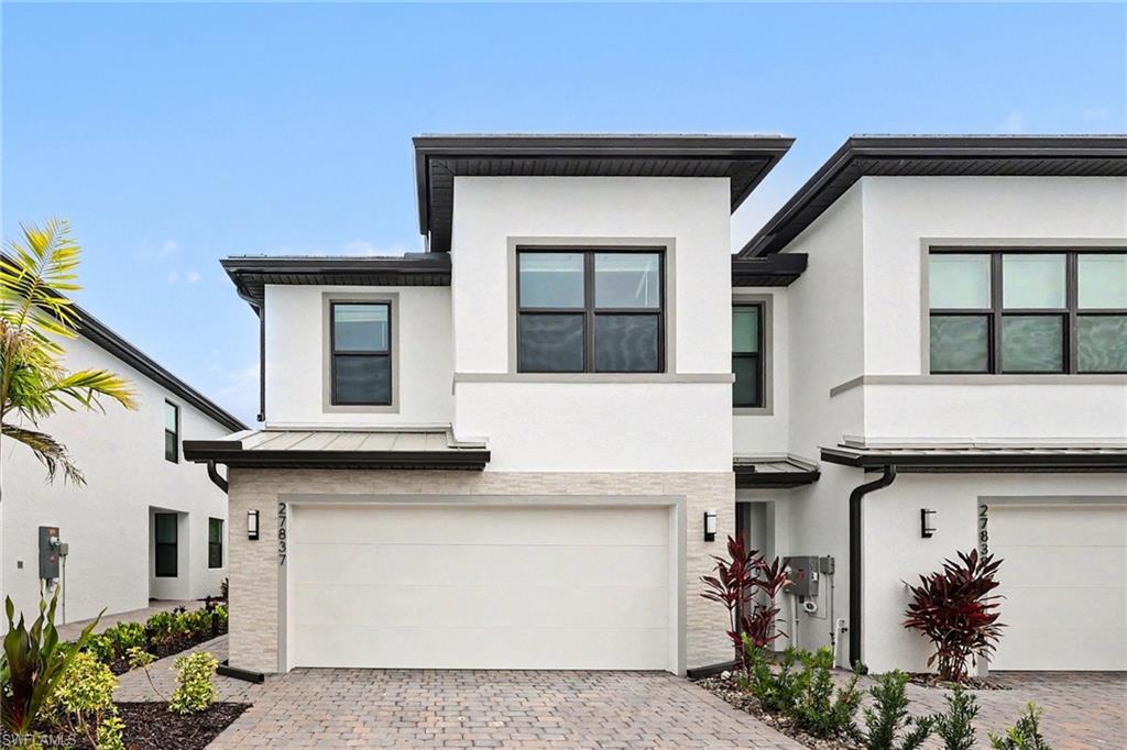 Contemporary townhouse exterior featuring white stucco, black-framed windows, a garage with a paver driveway, light-colored stacked stone accents, and dark soffit trim
