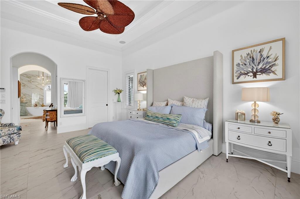 Bedroom featuring light marble finish flooring, arched walkways, a tray ceiling, a ceiling fan, and ornamental molding