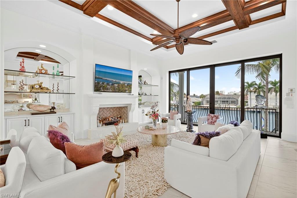 Living area featuring a fireplace with raised hearth, coffered ceiling, built in shelves, beamed ceiling, and ceiling fan