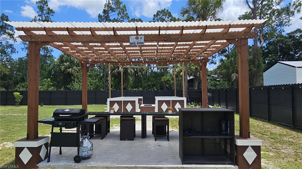 Outdoor entertainment area featuring a wood pergola with a translucent roof, a concrete pad, built-in counter spaces with diamond-patterned bases, and surrounding dark fencing