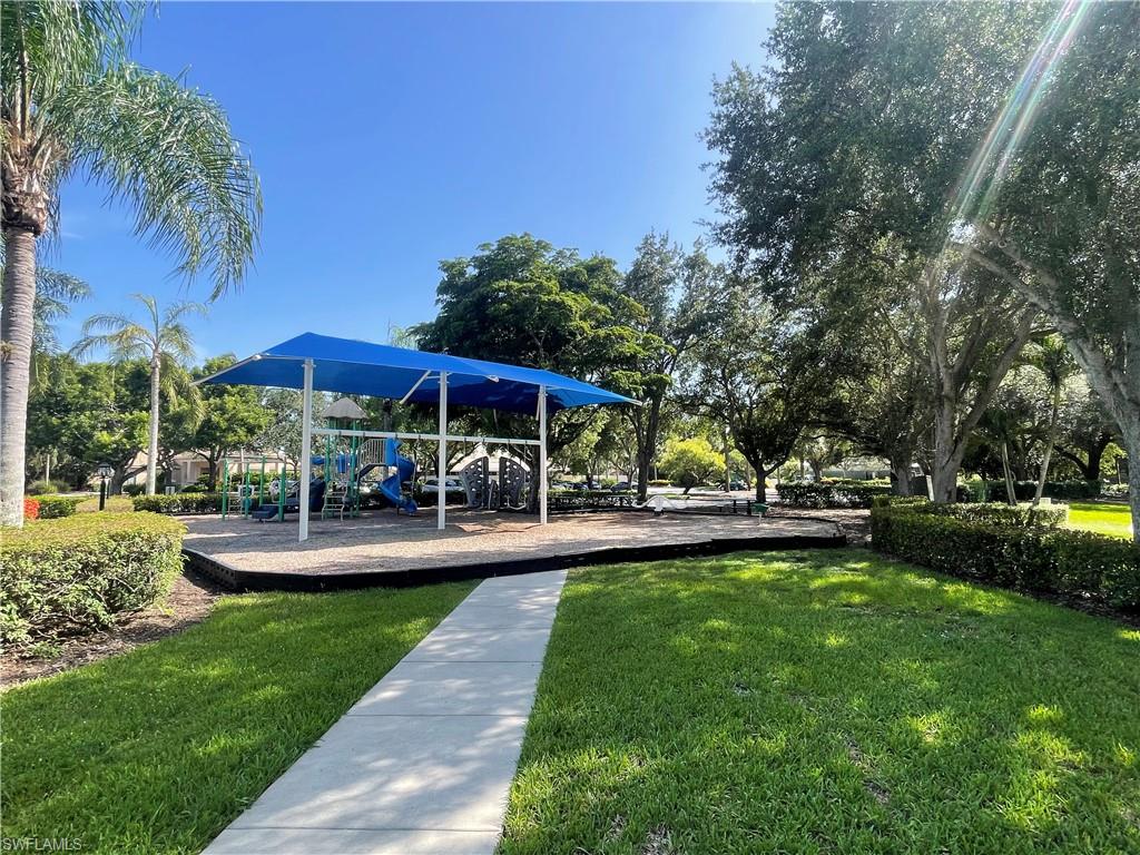 Community playground featuring a blue shade canopy, integrated play structures, and a surrounding hedge.