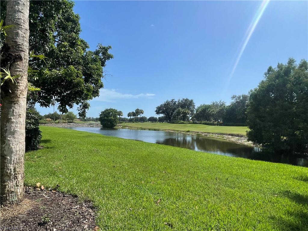 Expansive green lawn bordering a tranquil body of water, with mature trees and distant foliage under a clear sky.