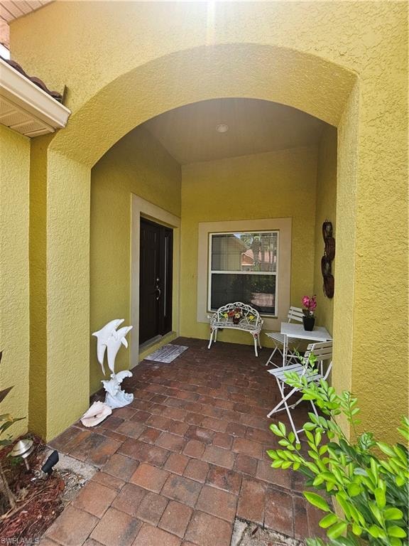 Arched entry porch featuring textured stucco walls, paver flooring, a dark entry door, and an exterior window with white trim